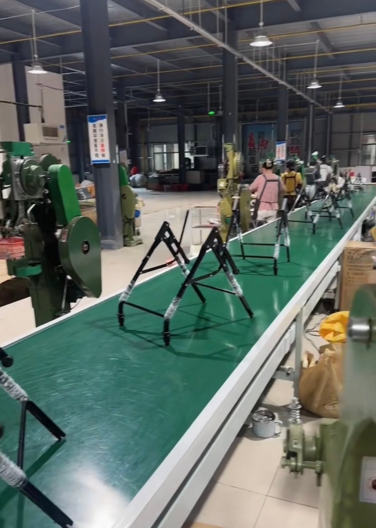 A close-up industrial photograph focused on a baby stroller manufacturing assembly line. A human factory worker, wearing safety glasses, stands at a dedicated riveting machine positioned alongside a moving conveyor belt. The worker is focused on joining stroller frame components (metal tubes and brackets), while multiple pre-assembled stroller bases move along the adjacent conveyor line, illustrating large-scale production.